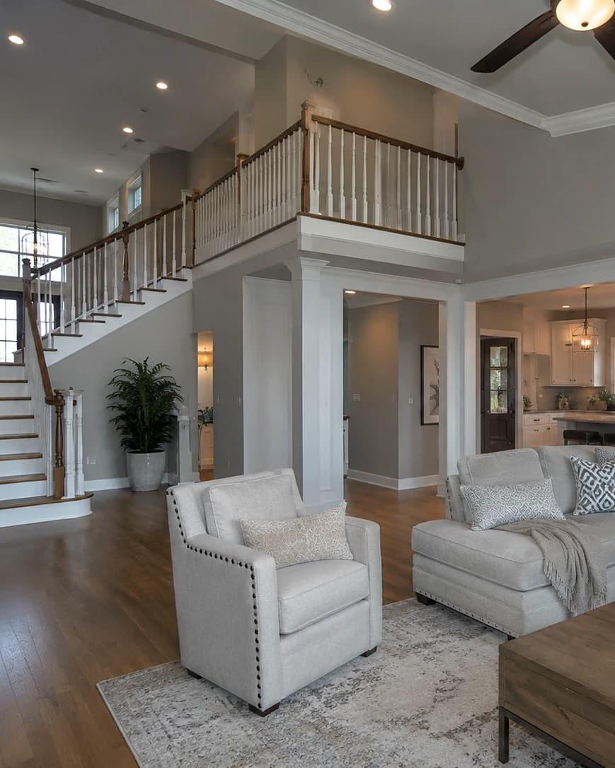 Luxury open-concept living room with double-height ceilings, a hardwood staircase, gray walls, and a white open kitchen beyond — high-end residential remodeling.