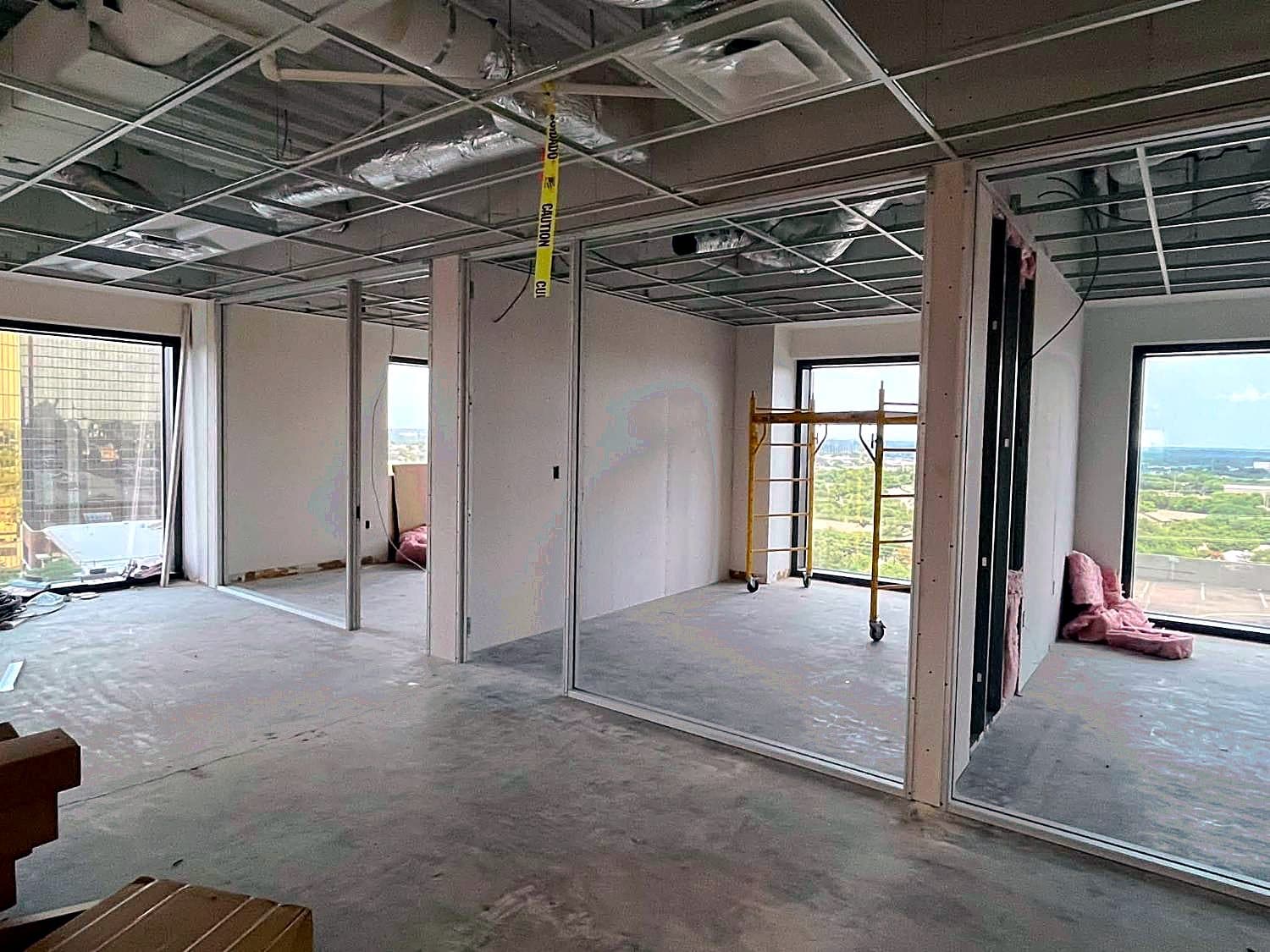 Open commercial office space framed with metal studs by REVIBE REMODELING, with the suspended ceiling grid installed and floor-to-ceiling windows beyond.