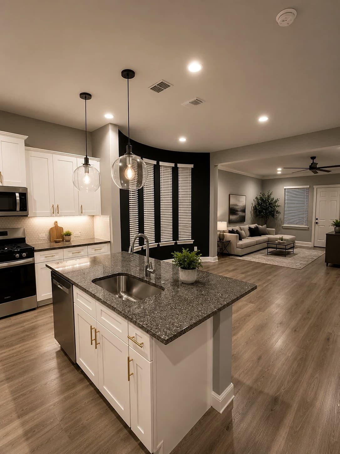 Open-concept kitchen and living area — white shaker cabinets, a granite-topped island with an undermount sink, glass globe pendant lights, stainless appliances, and a bright living room beyond.