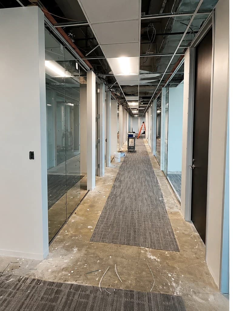 Commercial office corridor mid-build-out — open ceiling, bare concrete floor, and glass partition walls being installed.