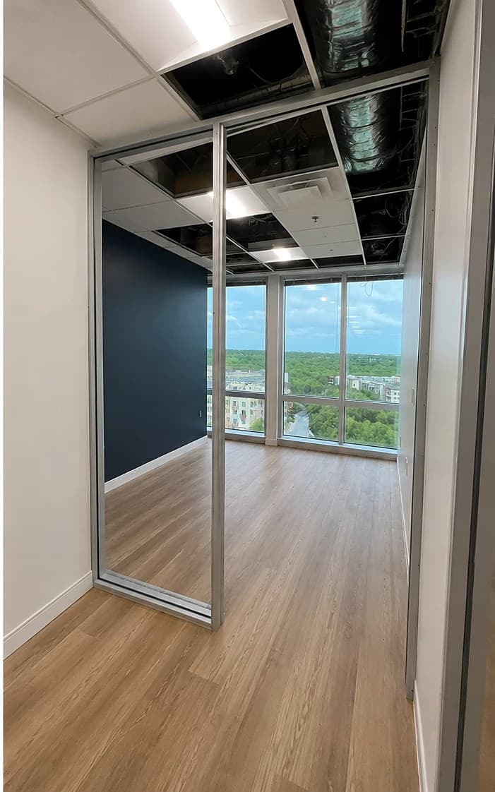 The same office finished — a glass partition wall, wood-look floors, a navy accent wall, and floor-to-ceiling windows with a city view.