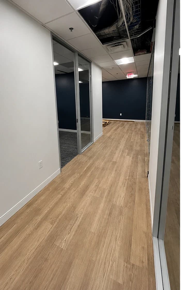 Private office mid-build-out — glass partition frames, an open ceiling section, a navy accent wall, and newly laid wood-look flooring.