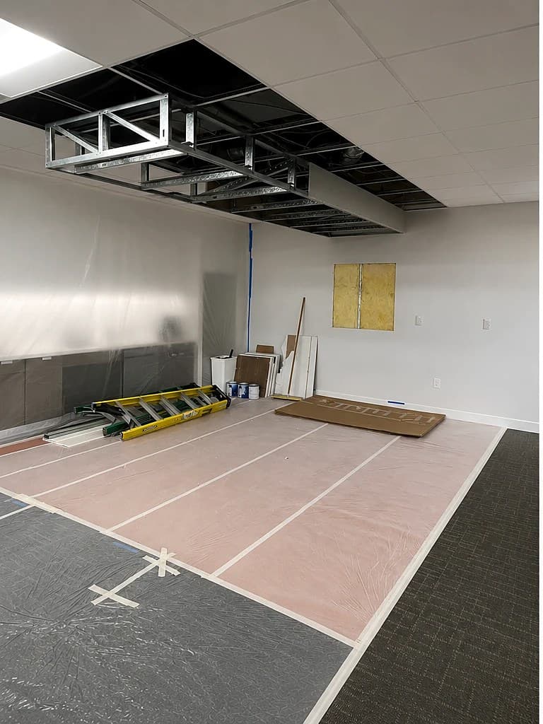 Commercial reception area mid-build-out — open ceiling grid, walls masked with plastic, and materials staged on a protected floor.